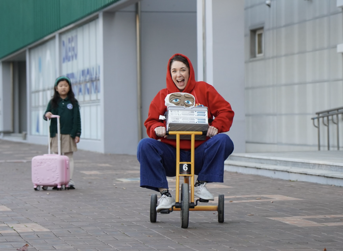 Mrs. Gum, impersonating E.T.,  excitingly pedals her tricycle. Her basket contains all of her materials for work.